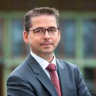 Mann mit kurzen dunklen Haaren und schwarzer, eckiger Brille. Trägt ein dunkelgraues Sacko mit weißem Hemd und roter Krawatte. Im Hinterrgund ist verschwommen ein Gebäude mit grünen Fensterrahmen zu erkennen.