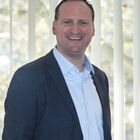 Mann mit kurzen, dunkeln Haaren, der lächelt. Trägt ein dunkelblaues Sacko, dazu ein hellblaues Hemd. Steht vor einem Fenster.