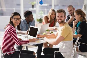 Zwei junge Erwachsene, die lächelnd in die Kamera schauen. Sie sitzen gemeinsam mit Anderen vor ihren Laptops an einem Tisch und tauschen sich aus.