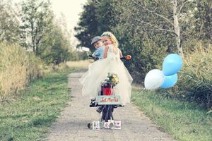 Zwei Kinder auf einem elektrischen Fahhrad. Der Junge sitzt am Steuer, während das Mädchen im Brautkleid hinten draufsitzt. Hinten am Fahhrad ein Schild mit der Aufschrift "Just Married", Dosen, die mit Fäden am Fahrrad befestigt sind und drei Luftballons