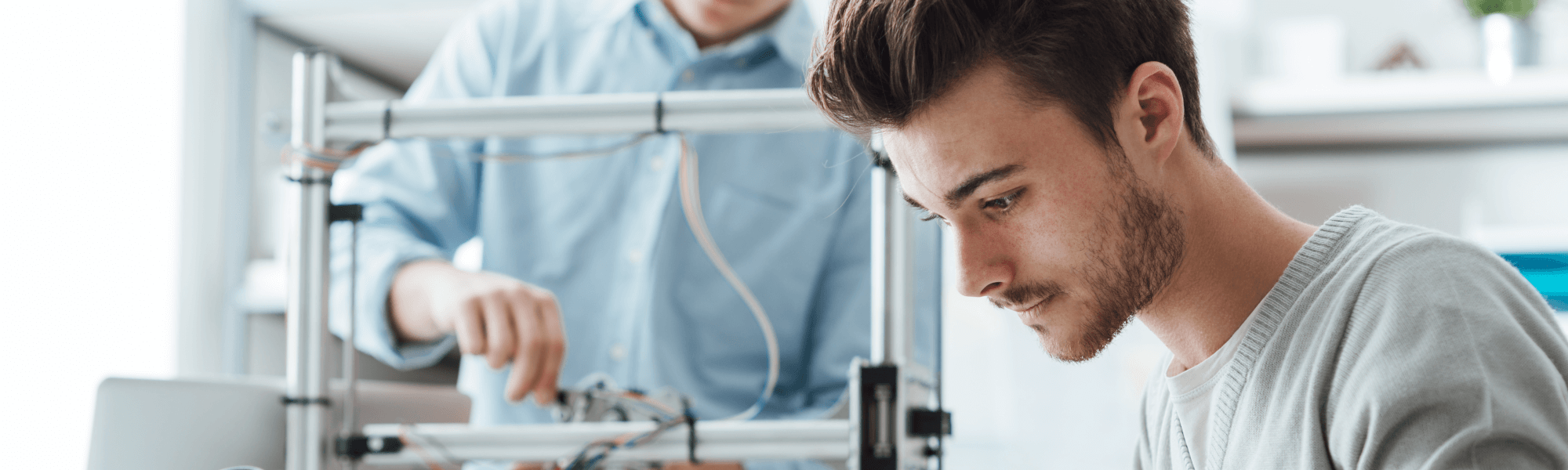 Rechts im Vordergrund ein Mann mit dunkelbrauner Kurzhaarfrisur und Dreitagebart. Schaut nach unten. Im Hintergrund ein Mann mit hellblauem Hemd, der an einem technischen Gerät arbeitet.