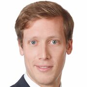 Mann mit rötlichen kurzen Haaren, lächelnd. In schwarzem Sacko und weißem Hemd. Der Hintergrund ist weiß.
