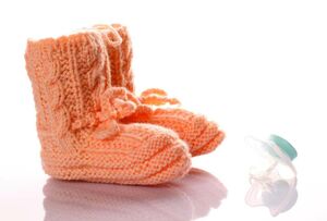 Lachsfarbene gestrickte Babystiefel. Rechts daneben ein Schnuller.