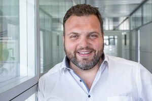 Mann mit kurzen dunklen Haaren und Bart, lächelnd. Trägt ein weißes Hemd. Im Hintergrund der Gang eines Gebäudes mit vielen Fenstern.