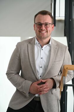 Mann mit hellbraunem kurzen Haar und leichtem Bart. Lächelt und mit ründlicher dunkler Brille. Trägt ein beiges Sacko und ein weißes Hemd.