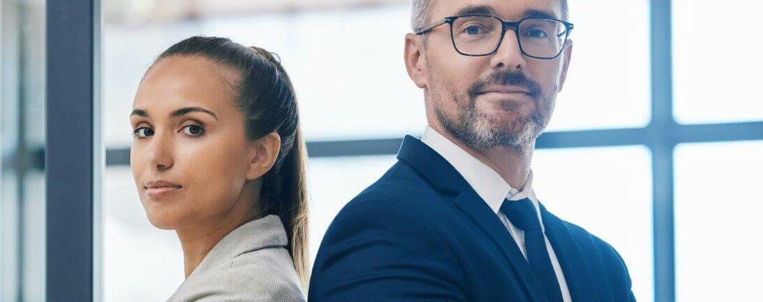 Mann und Frau im Business-Look, die mit verschränkten Armen Rücken an Rücken stehen und in die Kamera schauen.