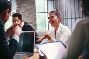 Frau im Business-Look, die ihren Gesprächpartner lachend anschaut und einhändig gestikuliert. Ihr Geprächspartner und der Mann neben ihr tragen schwarze Anzüge und sitzen vor Laptops.
