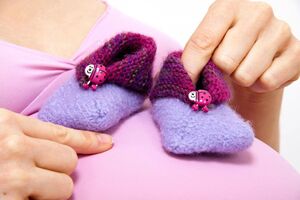 lila-rosa gestrickte Babysocken mit einem Marienkäfer, die zwei Frauenhände vor einen schwangeren Bauch halten.