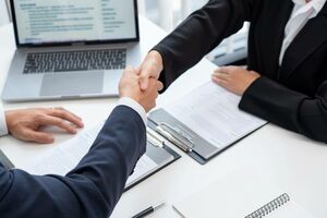 Der Handschlag zweier in Blazer/Sacko gekleideter Personen über einem Schreibtisch. Auf dem Schreibtisch vor ihnen ein Laptop und je ein Klemmbrett.
