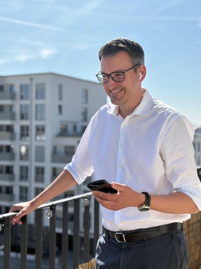 Mann mit dunkler Kurzhaarfrisur und schwarzer, eckiger Brille, lächelnd. Trägt ein weißes, hochgekrämpeltes Hemd, eine schwarze Armbanduhr und kabellose Kopfhörer. Er hält ein Handy in der Hand. Steht auf einem Balkon, im Hintergrund andere Häuser.