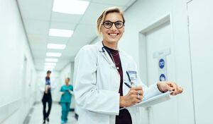Junge Ärztin, die mit einem Klemmbrett unter dem Arm im Gang eines Krankenhauses steht und sich Notizen macht.
