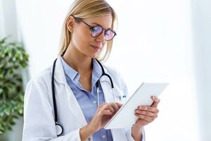 Junge Ärztin im weißen Kittel und mit Stethoskop um den Hals, die ein Tablet in den Händen hält.