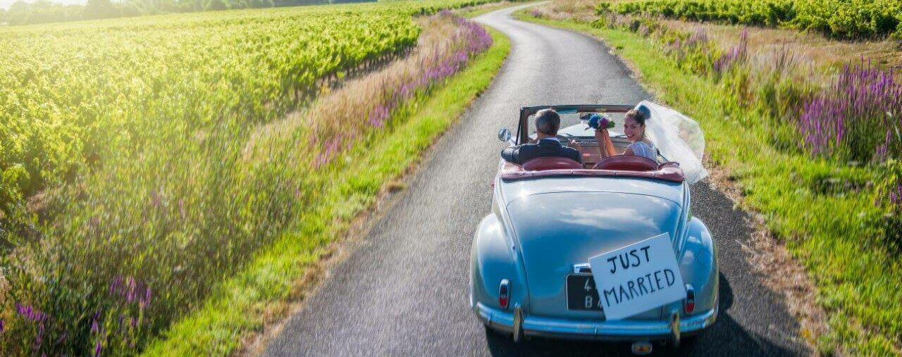 Frisch verheiratetes Ehepaar, die in einem alten Auto einen Feldweg entlang fahren. Hinten am Auto ein Schild mit der Aufschrift "Just Married".