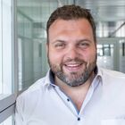 Mann mit kurzen dunklen Haaren und Bart, lächelnd. Trägt ein weißes Hemd. Im Hintergrund der Gang eines Gebäudes mit vielen Fenstern.