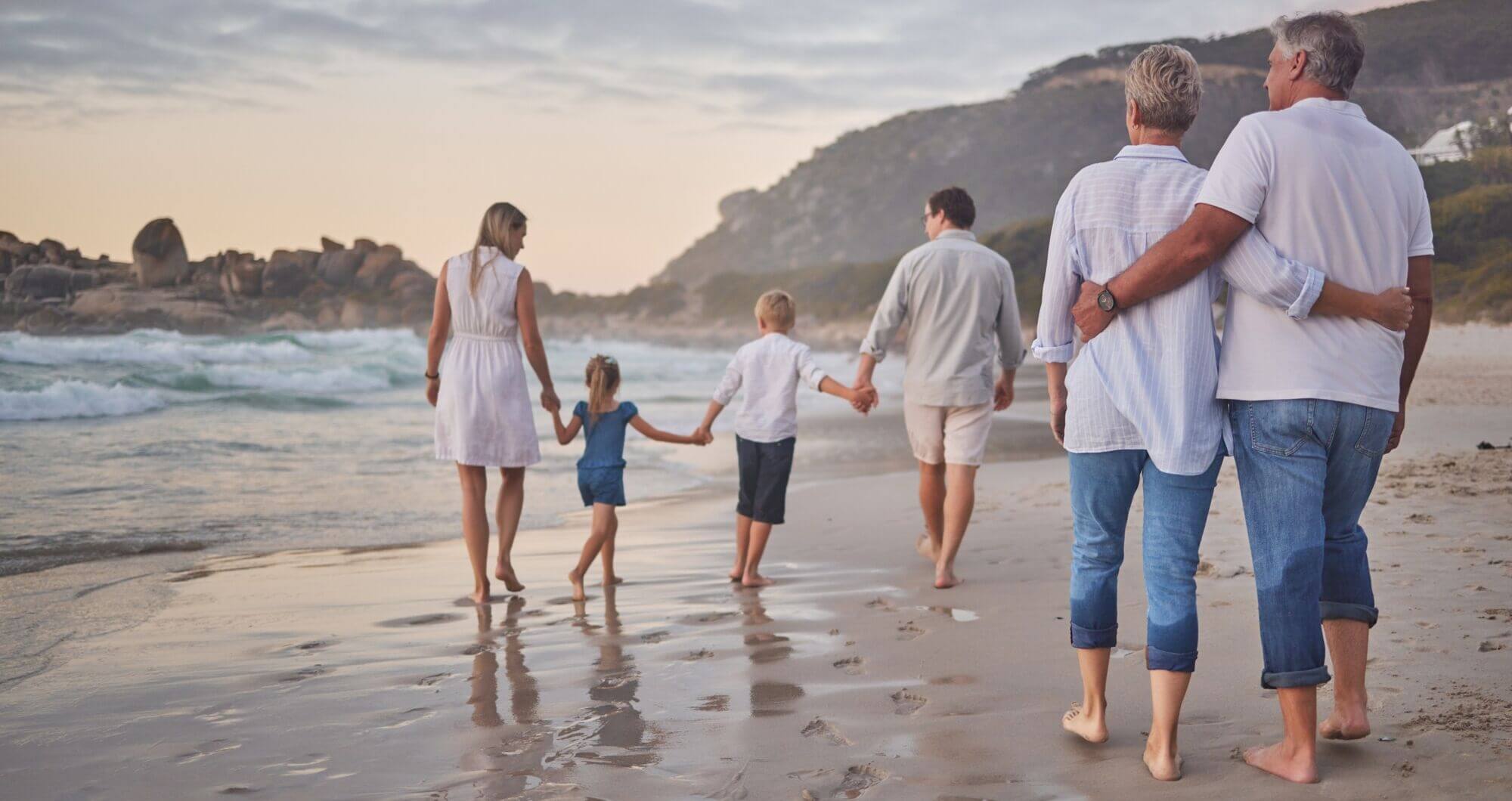 Rechts ein Mann und eine Frau mit grauen Kurzhaarfrisuren, die barfuß Arm in Arm laufen. Links vor ihnen eine vierköpfige Familie, die in einer Reihe Hand an Hand barfuß im Sand laufen. Alle sind von hinten zu sehen. Links das Meer, im Hintergrund Berge.