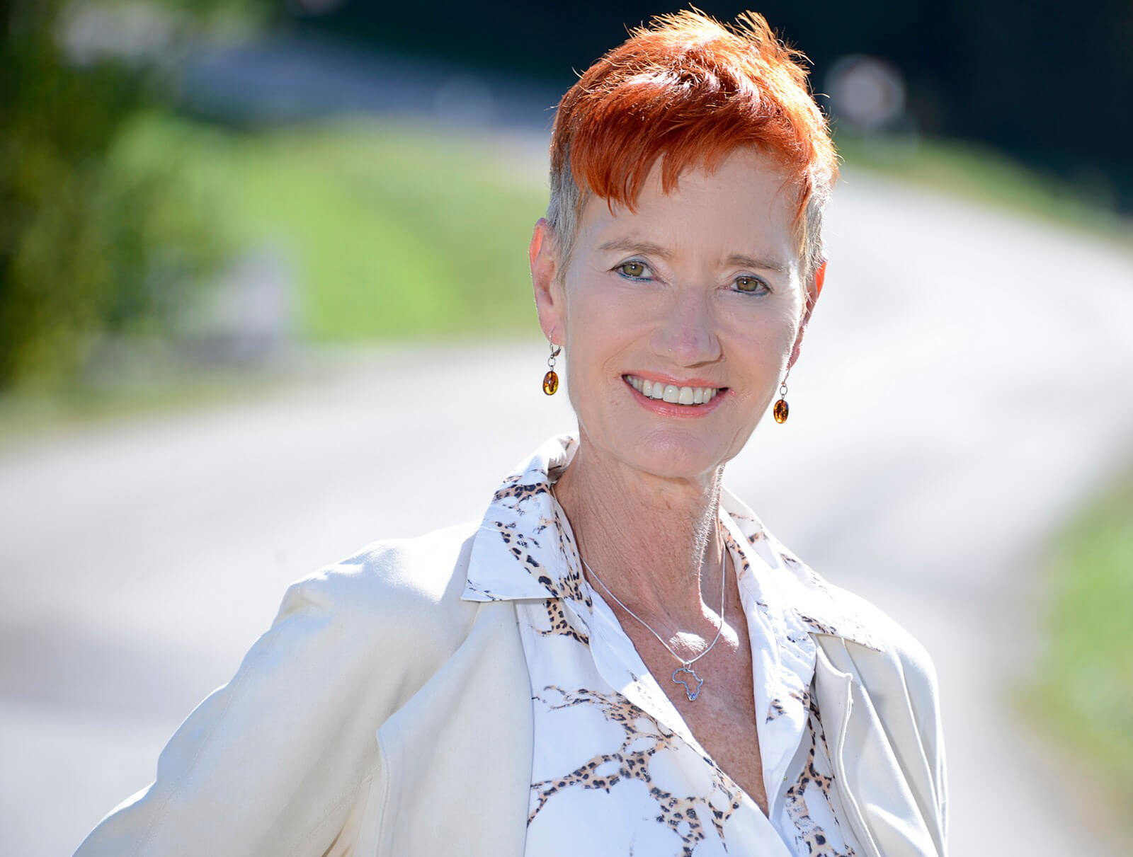Frau mit kurzen roten Haaren, lächelnd. Trägt einen beigen Blazer und eine gemusterte, weiße Bluse. Im Hintergrund ein Schotterweg und Wiese.